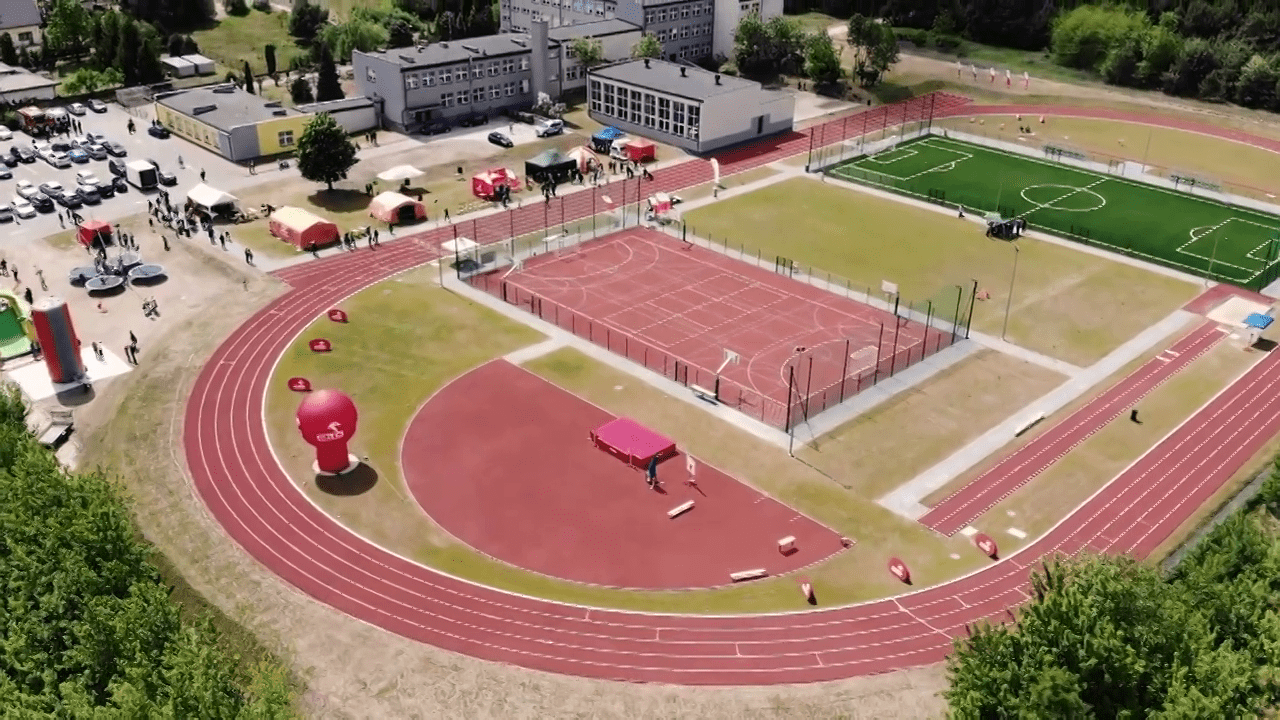 Zdjęcie przedstawia widok z lotu ptaka na kompleks lekkoatletyczny przy szkole podstawowej w Młodzieszynie. Na terenie kompleksu widać poruszające się postacie. W oddali widać samochody na parkingu oraz namioty.